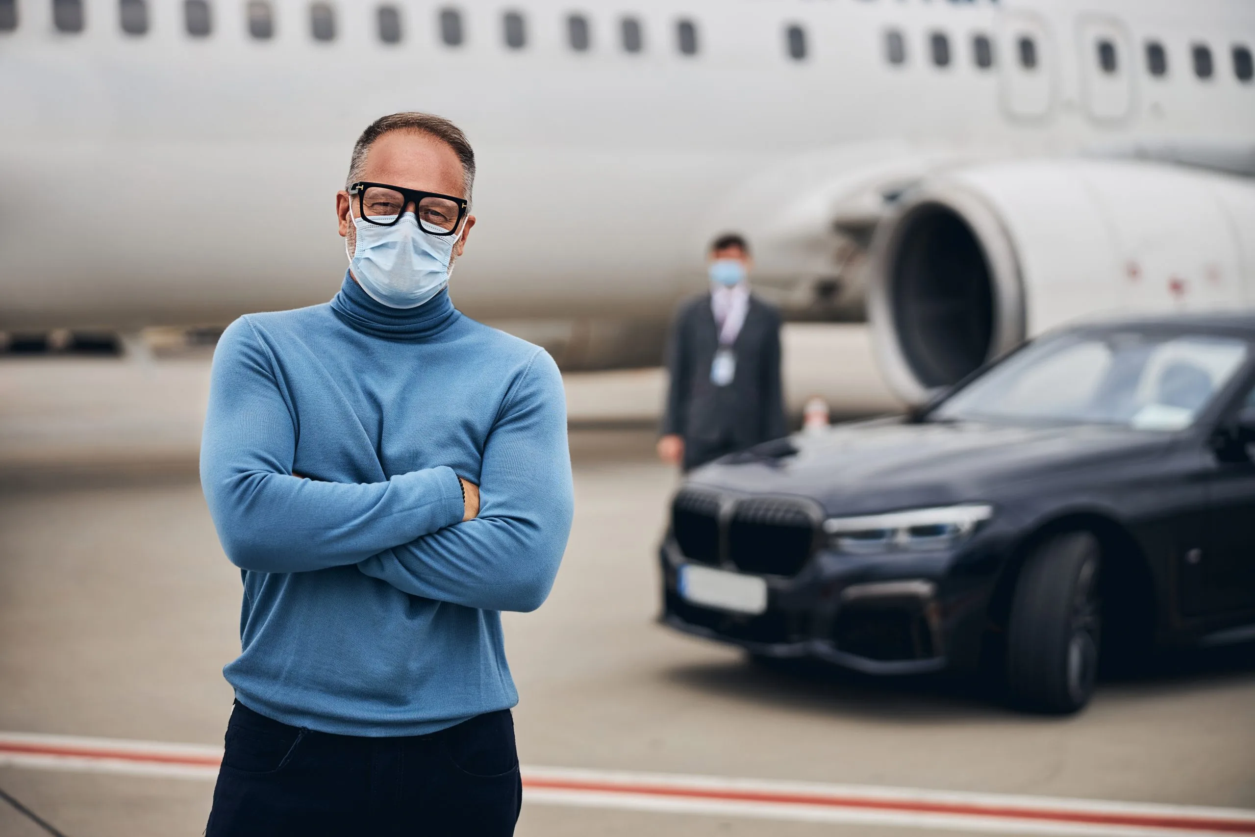 Airport Cabs - Passenger wearing a face mask and eyeglasses, staring ahead with a plane and luxury cab in the background, symbolizing safe, executive airport transport.