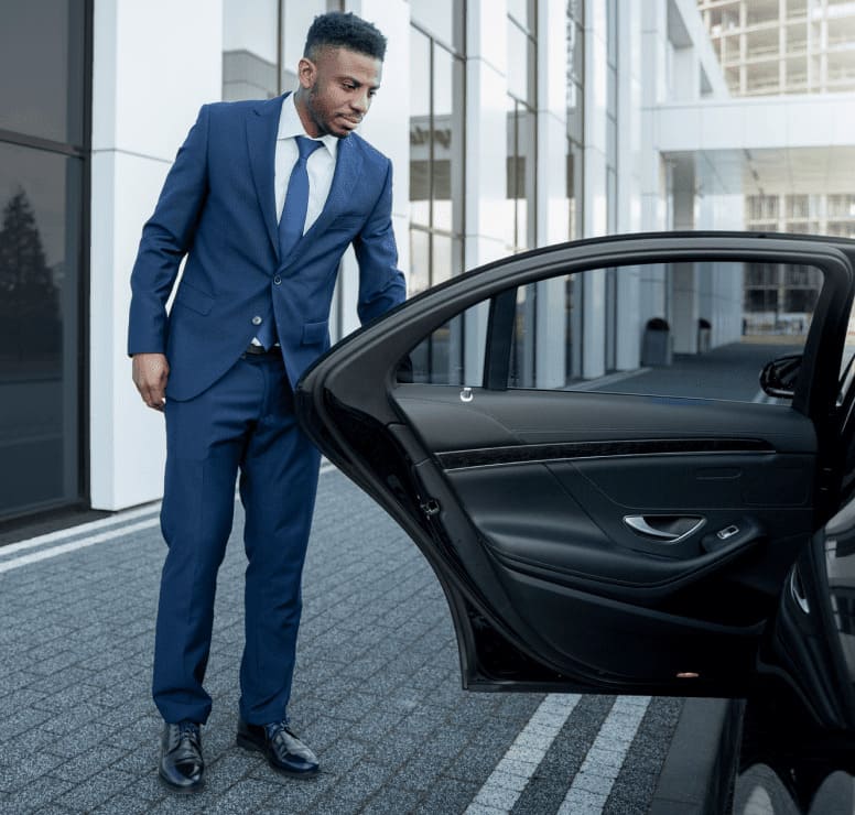 Professional chauffeur opening a luxury car door for an executive pickup outside a modern office building.​