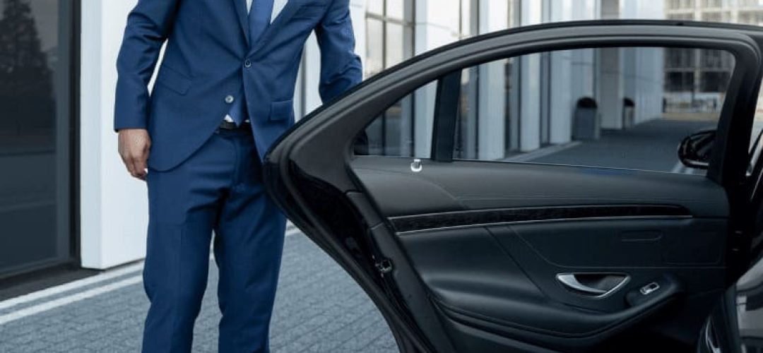 Professional chauffeur opening a luxury car door for an executive pickup outside a modern office building.​